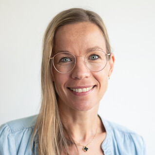 Frau mit blonden langen Haaren, runder Brille und einem Lächeln.